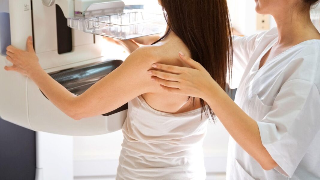 A generic picture of a woman having a mammogram. She is in front of a machine with another woman helping her.