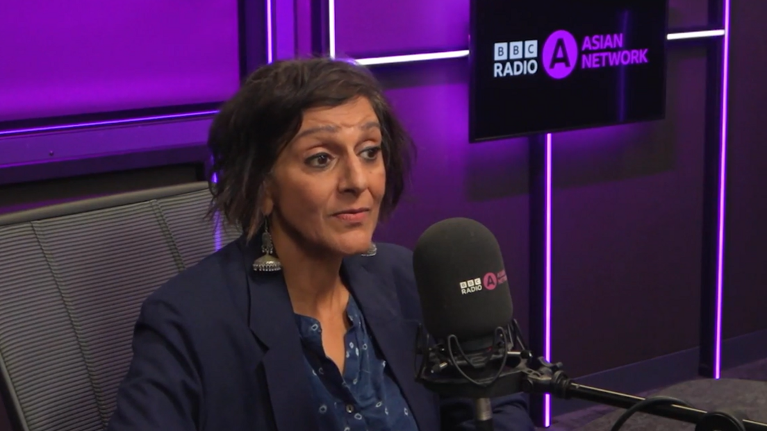 Dame Meera Syal a woman with chin-length brown hair and long silver earrings is wearing a navy blue blazer over a navy blue shirt with small white flowers on. She is sat behind the desk in a radio studio talking into a microphone. Behind her the wall is purple and on the right there is a black screen with the words BBC Radio Asian Network