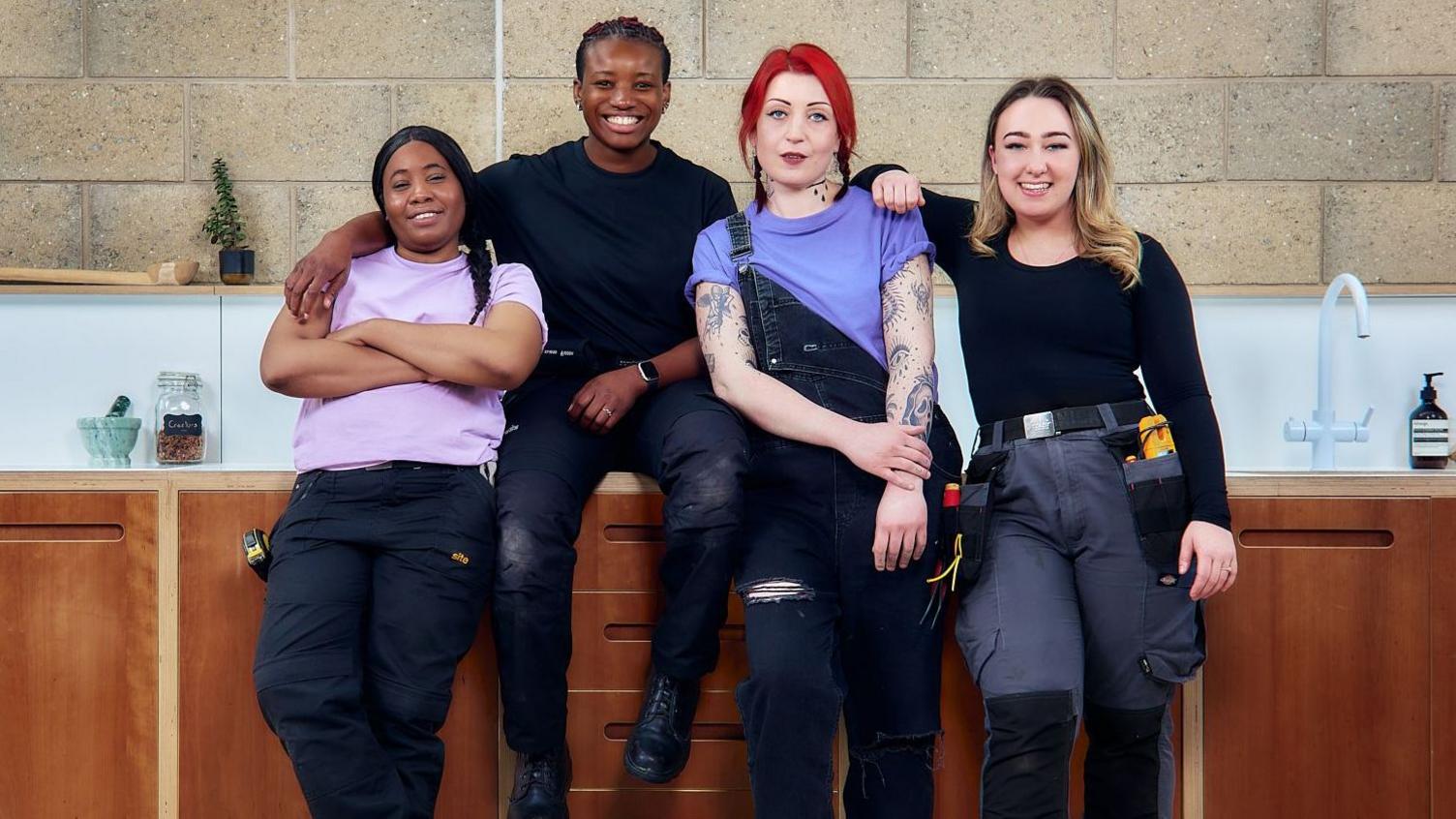 Four TaskHer tradeswomen smiling as they sit or lean against a kitchen worktop.