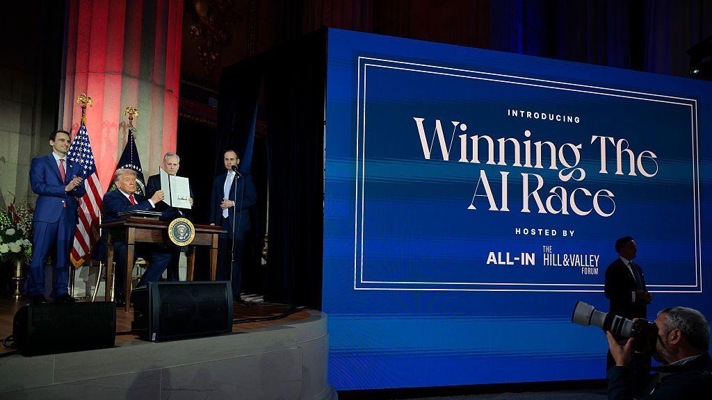 US president Trump, flanked by several members of his administration, sits behind a desk with the presidential seal on stage at an event in 2025. To the right of him is a huge blue sign that says "Winning the AI Race" in white font. He is holding a folder containing an executive order he had just signed on AI.