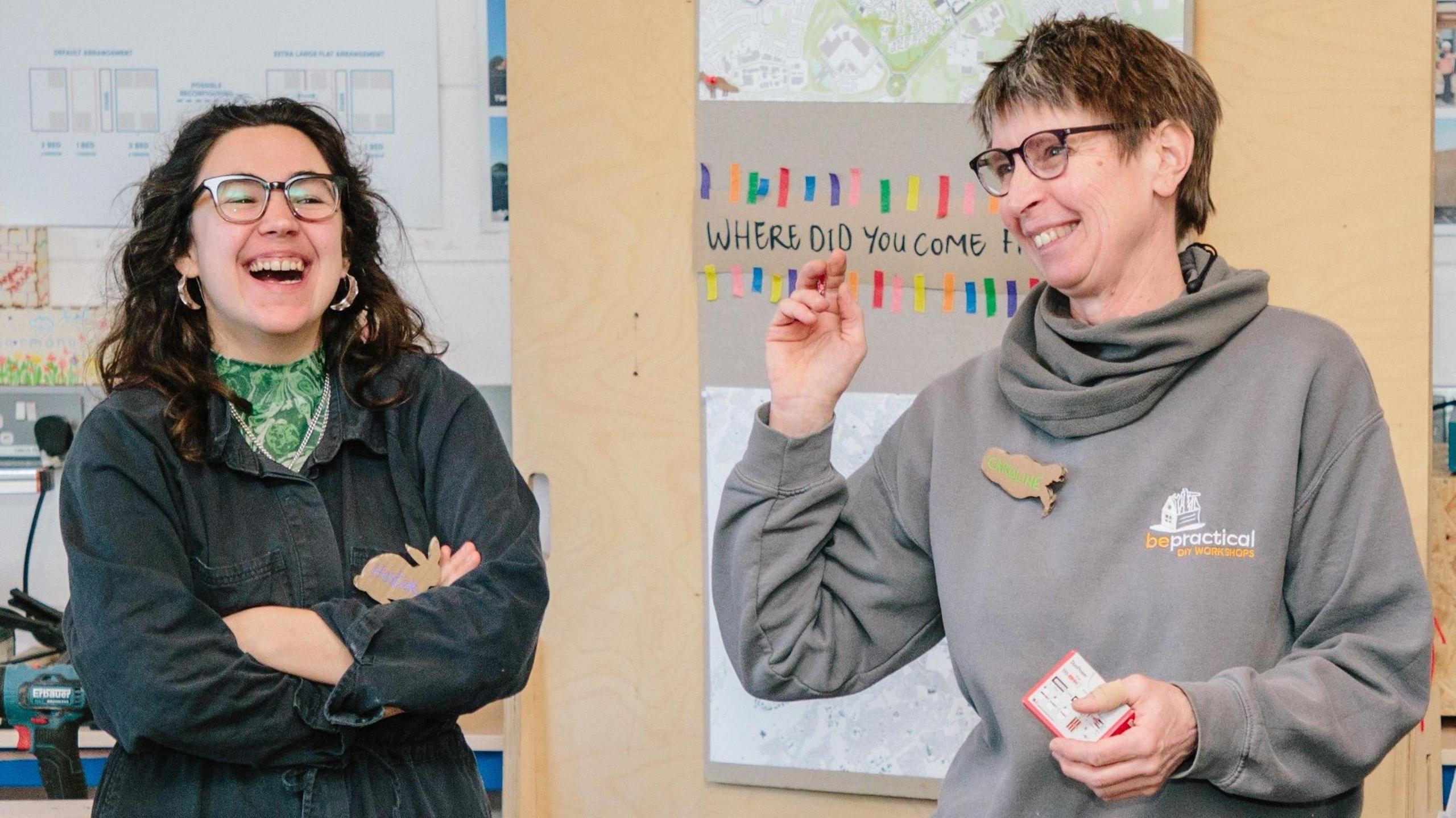Two women smile as they stand next to each other. One has long dark curly hair, is wearing glasses and has her arms folded. She is wearing a dark shirt with a green top underneath. The other woman is wearing a grey hoodie. She is wearing glasses and has short dark hair