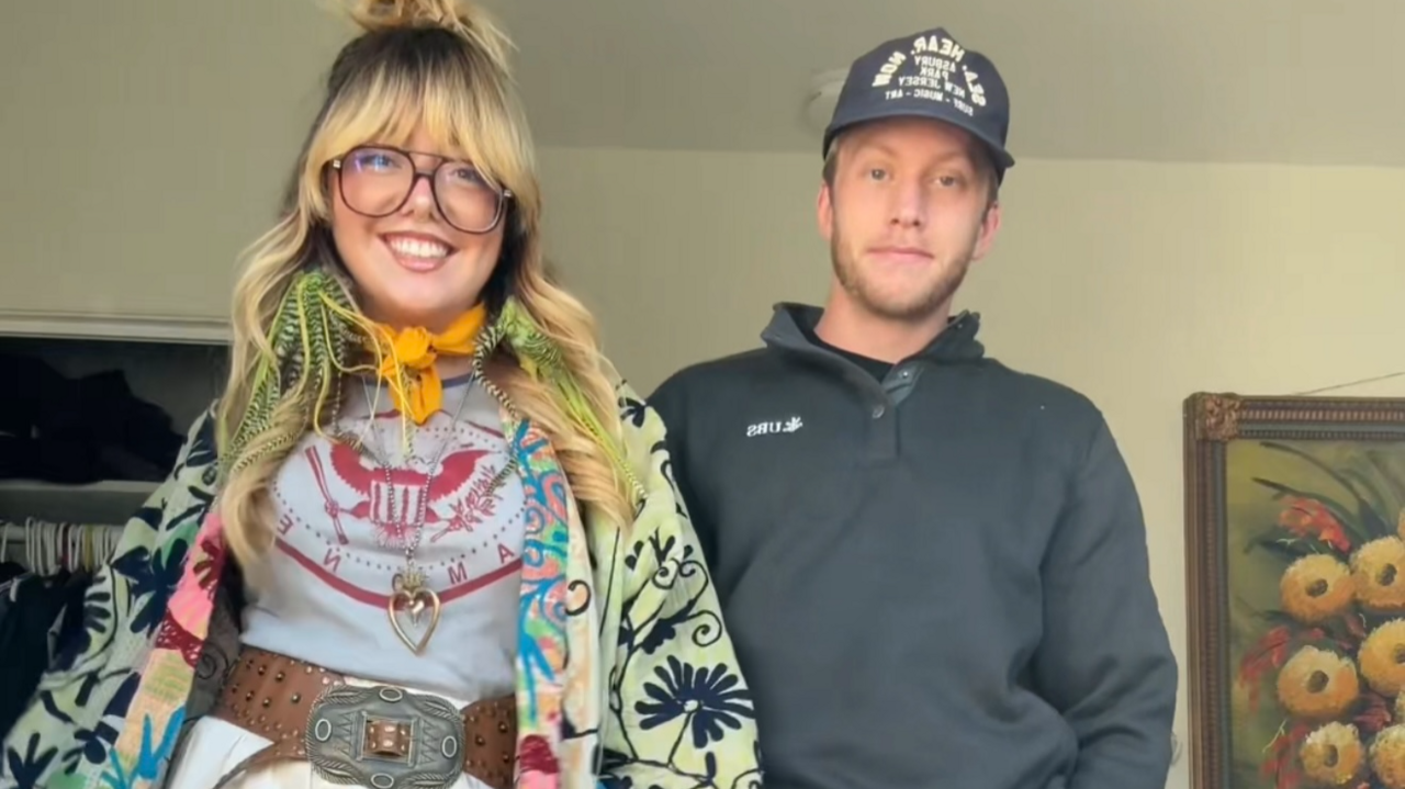 Jessica Raiola wearing a green and blue flower patterned jacket, orange neck scarf, grey and red t-shirt and a belt, next to her boyfriend wearing a dark cap and long-sleeved top.