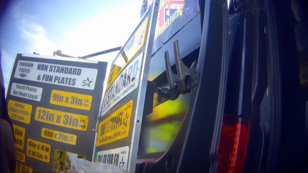 Display boards at a market stall advertising vehicle registration plates and related products. The boards show examples of standard plates, metal plates, plates for EU travel, tinted plates and other custom sizes, with pricing information visible. High-visibility clothing and market stalls can be seen in the background.