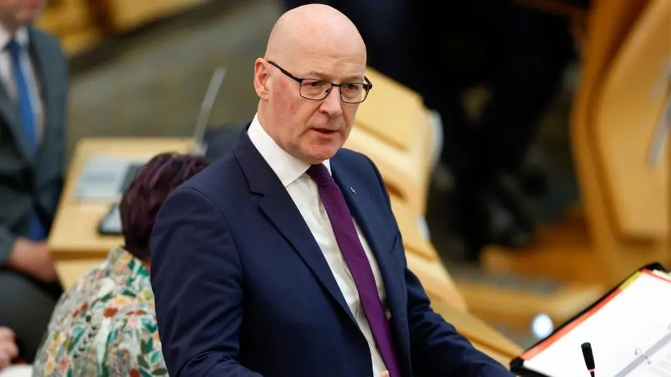 John Swinney, who is bald with glasses and wearing a navy business suit with purple tie, stands in a debating chamber.