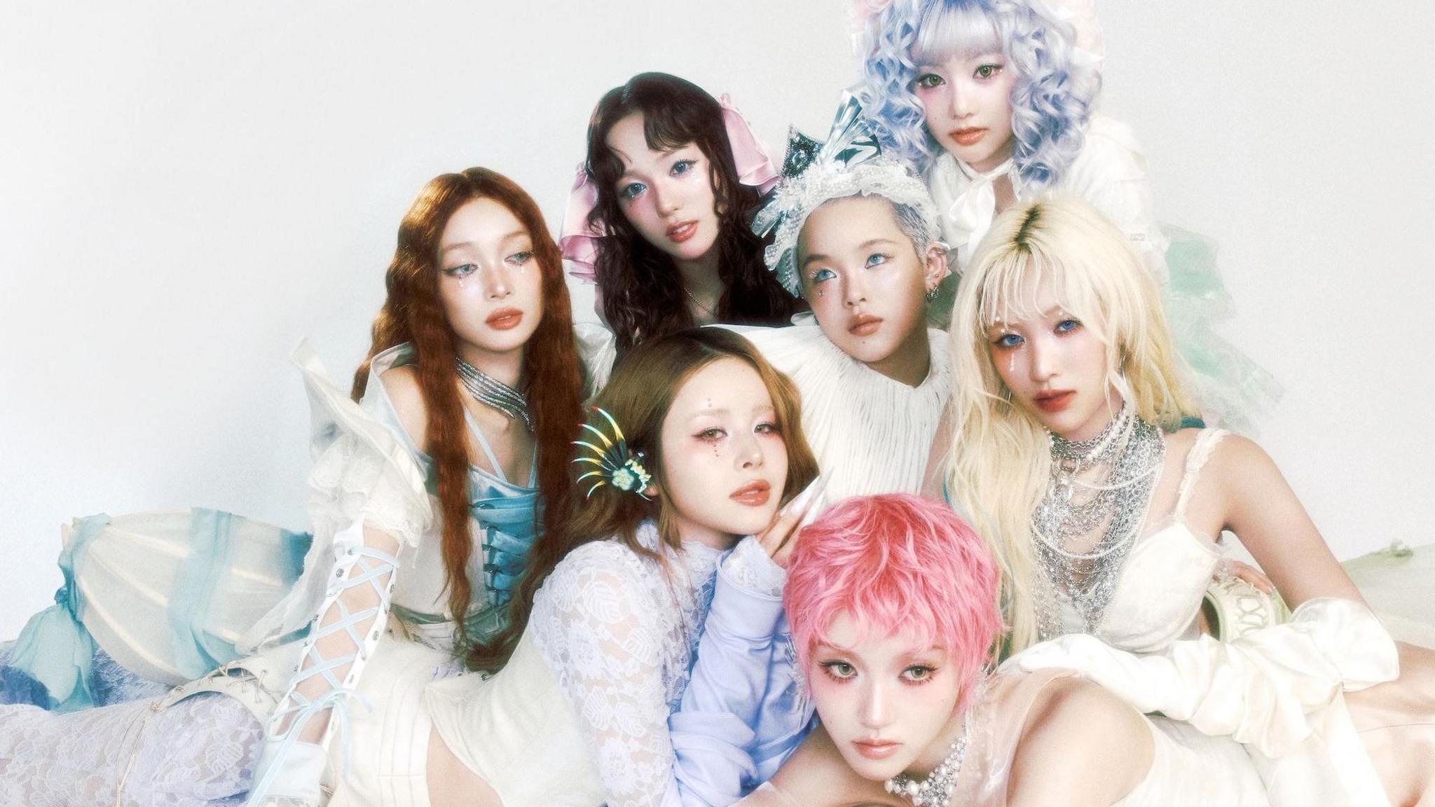 The seven members of XG pose for a promotional photo in a triangular formation, weaing a variety of pastel shaded, Victorian-inspired outfits