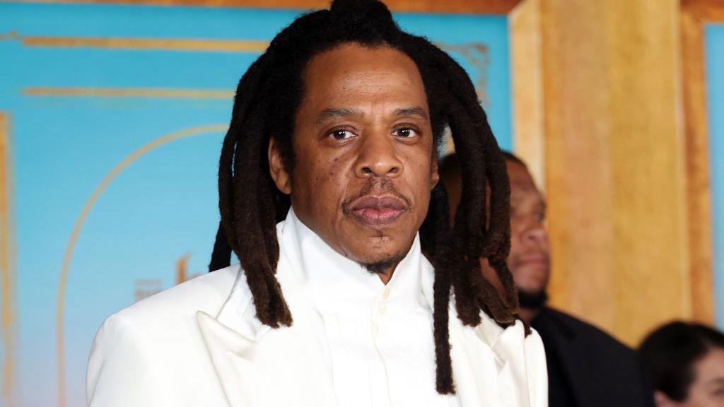 Jay-Z in a white suit at a party. He has a neutral facial expression.