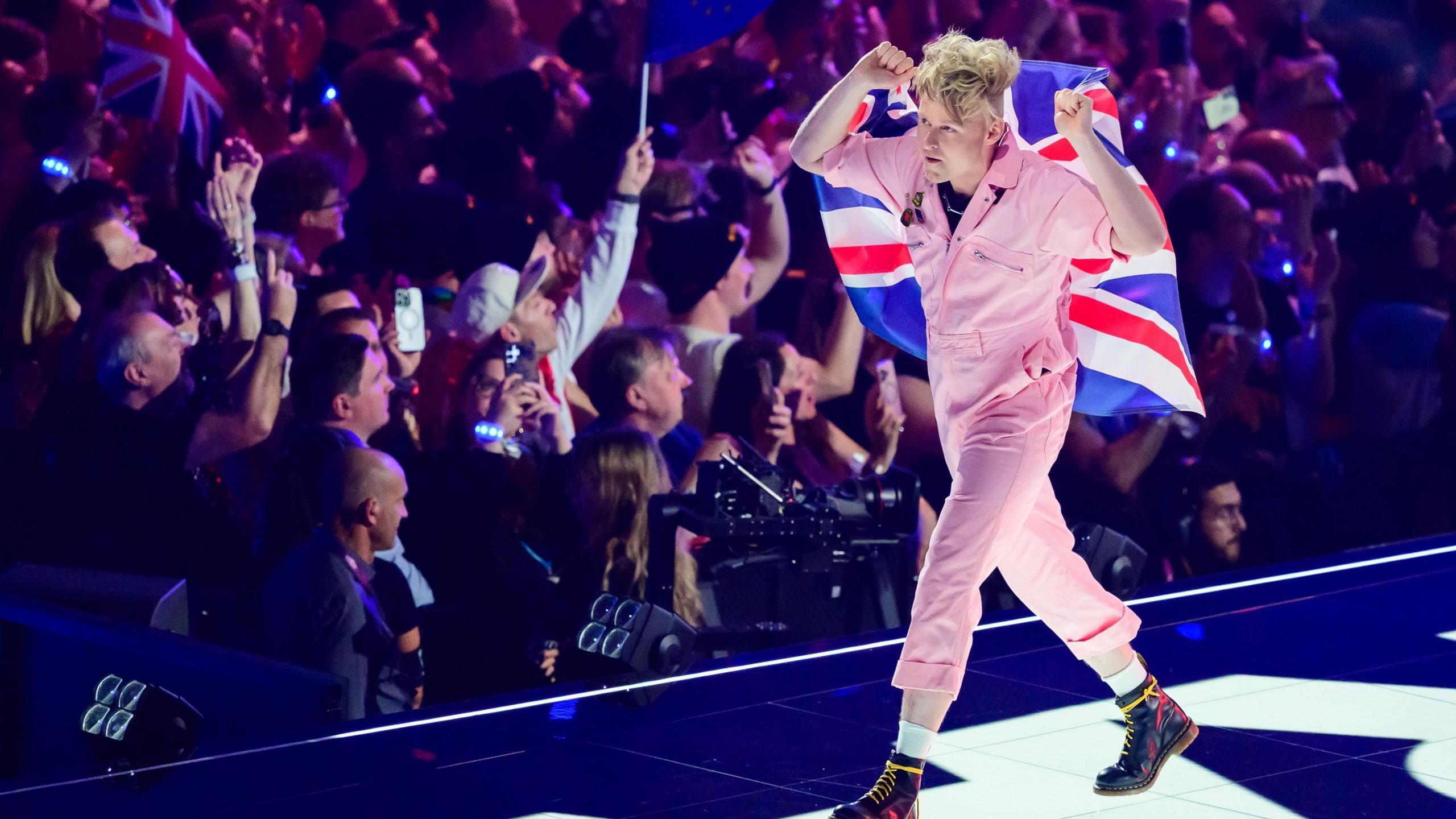A performer wearing a pale pink one-piece outfit walks along a brightly lit Eurovision stage draped in a large Union Jack flag. Audience members line both sides of the stage, many holding phones and flags. Purple and blue stage lights illuminate the arena and create a colourful backdrop around the performer.