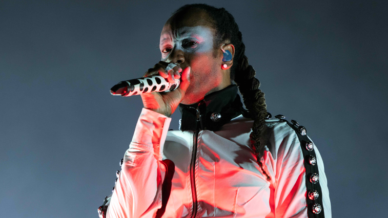 A man with a black and white tracksuit jacket top which has studs down the arms. He is singing into a black and white spotted microphone and has silver face paint around his eyes. He wears his hair in a long braid to the side.