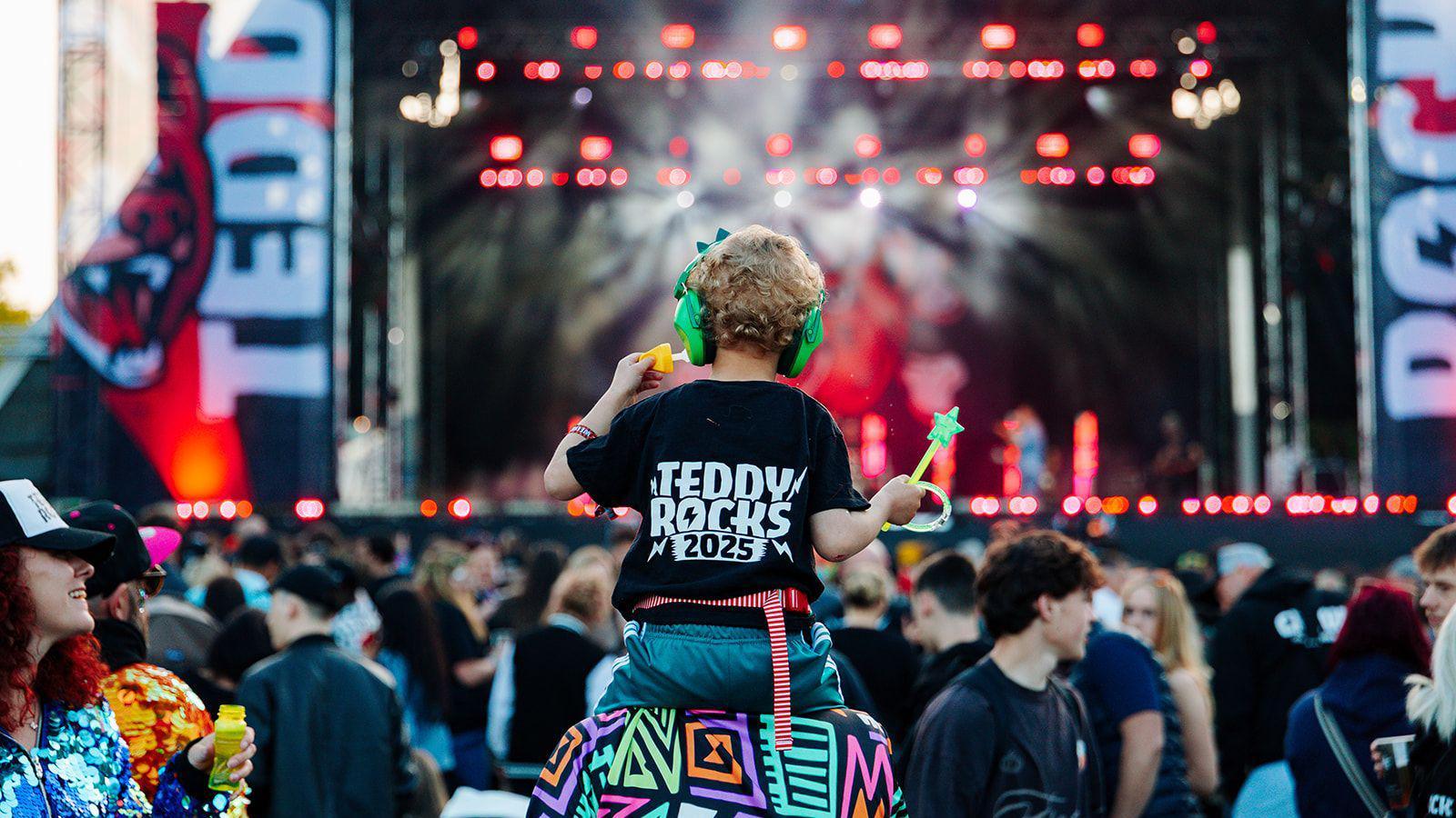 A crowd at Teddy Rocks festival in front of their stage. A child is seen on the shoulders of an adult in the middle of the crowd. The child is wearing a black shift reading Teddy Rocks 2025.
