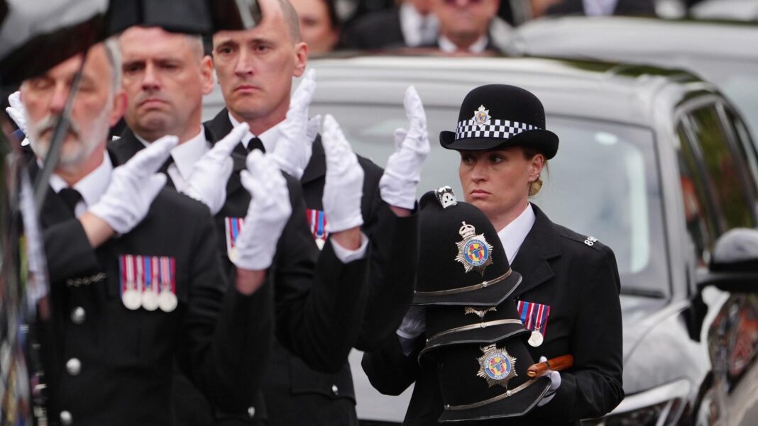 Four police officers with sad looks on their faces. Three of them are men who are standing in a line their hands over one of their shoulders with white gloves on. They are wearing black suits with three medals on their right chest. A female police officer in a round hat has collected the male officers' hats and is looking to the left. She has blonde hair which is in a bun. There is a black car behind them.