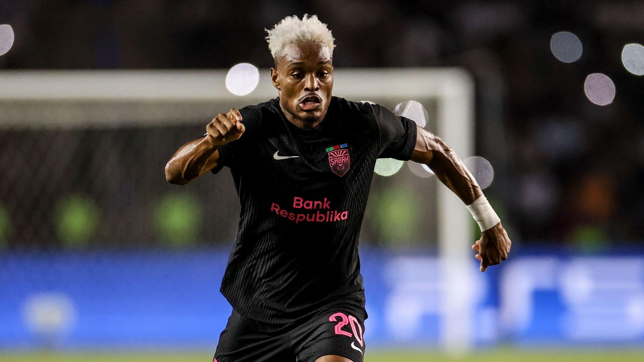 Joy-Lance Mickels, wearing an all-black Sabah kit with bright pink trim and number 20 on his shorts, spreads his arms wide as he is captured mid-stride during a football match. His hair is dyed white and he has a strange look of extreme effort on his face