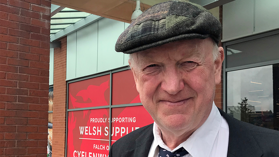 A close-up photograph shows a man wearing a dark patterned flat cap, black jacket, white shirt and dark tie, looking towards the camera in a near smile. He is outside a modern brick and glass building. Behind the person is a large red sign featuring a Welsh dragon