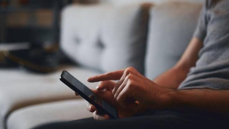 Close up of omeone sitting on a sofa holding a black phone, wearing a grey t-shirt