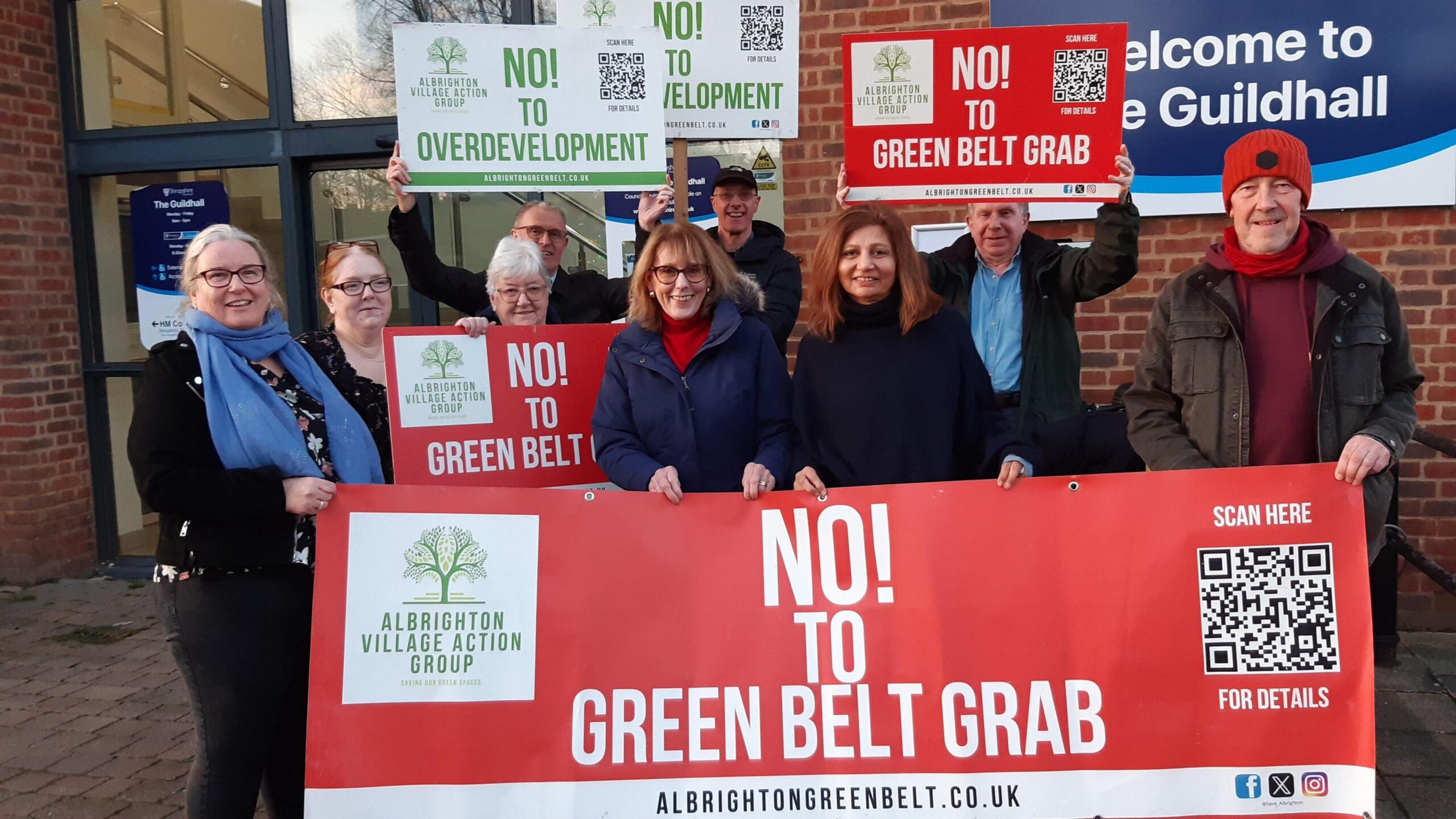 A group of people standing outside the Shropshire Council building that has a blue and white sign on the wall. They are smiling and celebrating, holding signs and boards that read "no to overdevelopment" and "no to green belt grab".