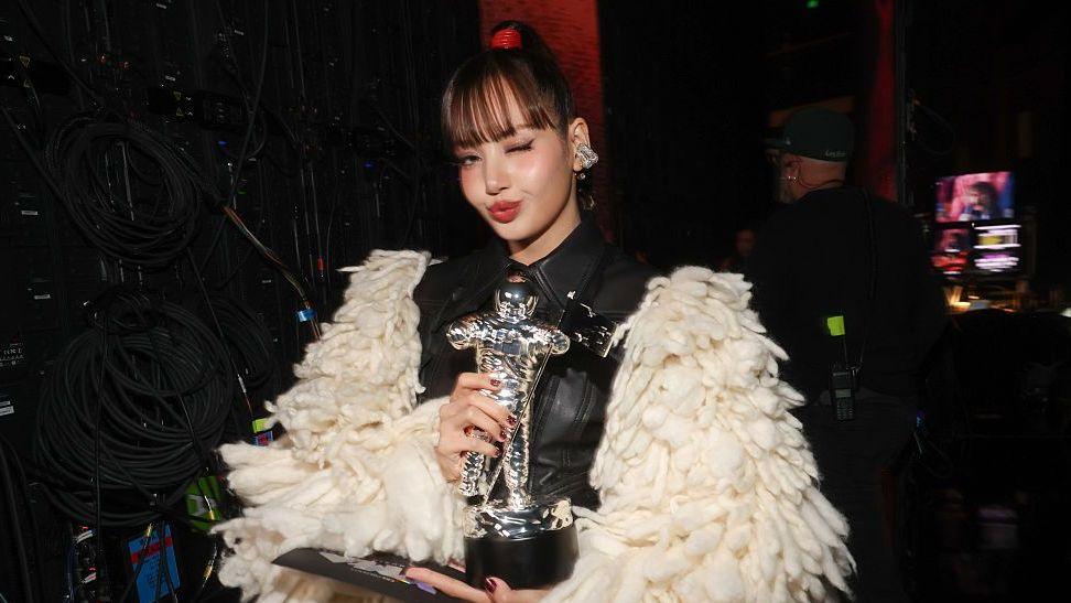 Lisa stands backstage in a dimly lit production area, holding a silver astronaut-shaped trophy and a flat award plaque. She wears a black outfit with an oversized cream-colored textured coat featuring thick, looped fabric. One eye is closed in a posed expression.