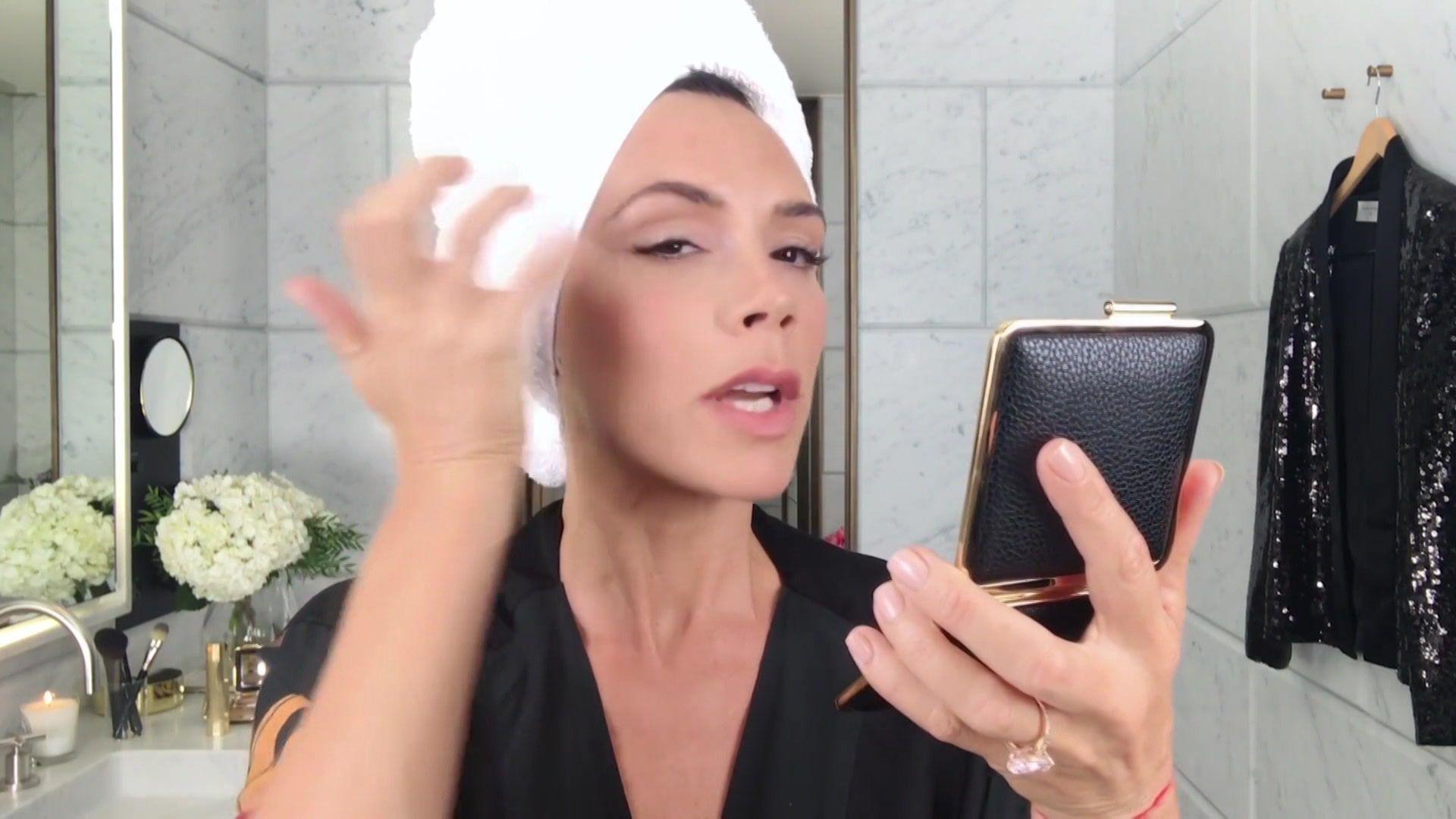 Victoria Beckham does her make-up in front of her bathroom mirror. She is wearing a white towel on her head and uses a powder compact
