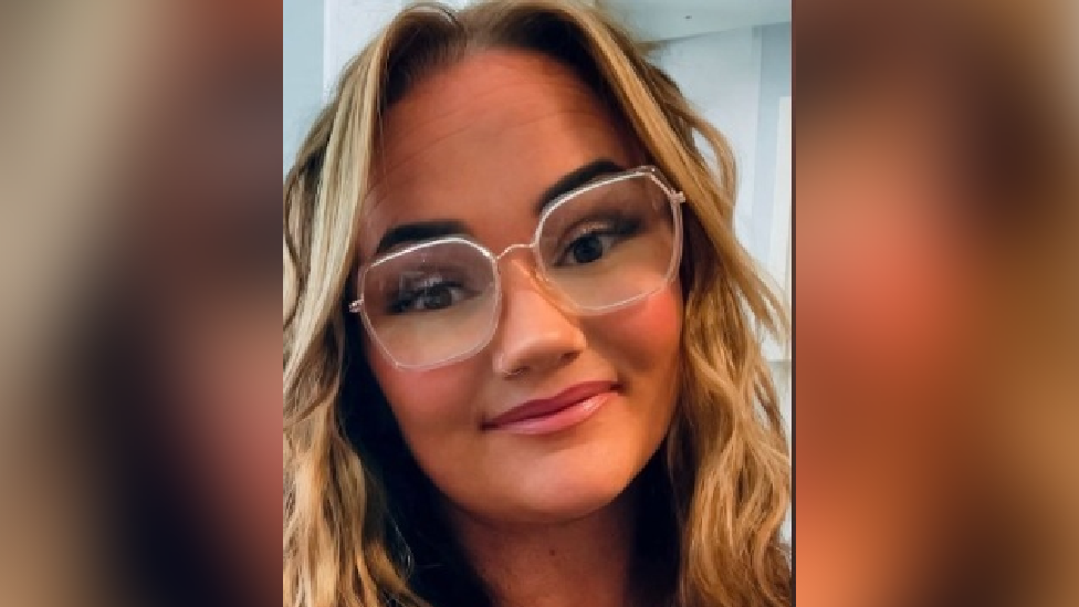 A blonde woman with a full face of makeup. Her hair is wavy and she's wearing clear big glasses, looking directly into the camera.