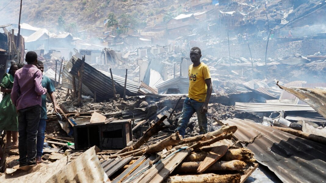 Man in yellow t-shirt stands in piles of rubble and corrugated iron while three other people stand on the left of the picture. The rubble seems to go on for some distance and the air is hazy.