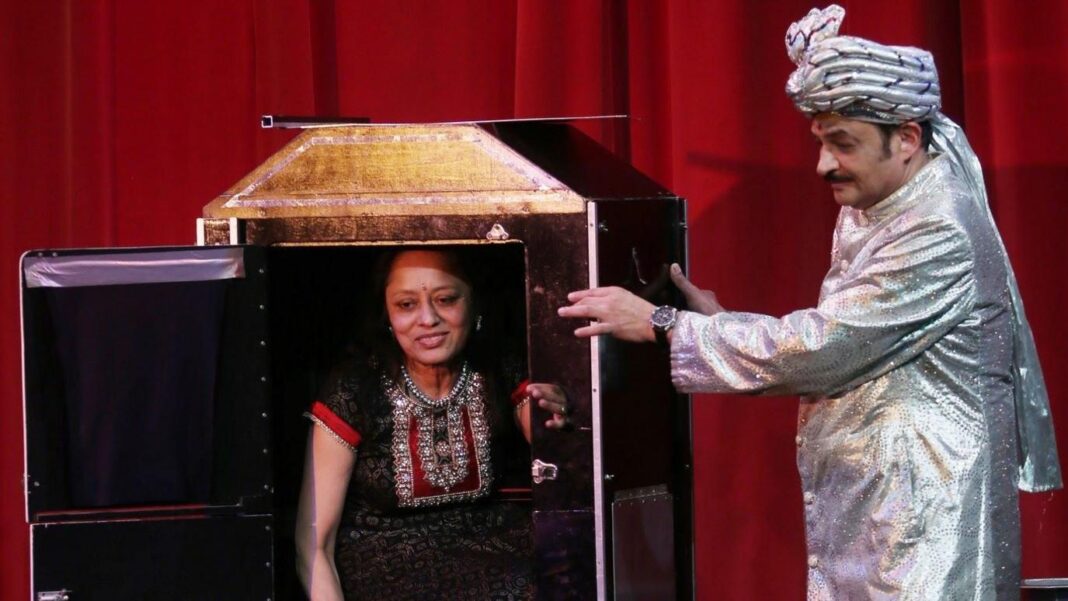 A hindu woman wearing a black and red sari is inside a box with a hinged door to the left and she is smiling. A man in a silver jacket with a silver pagri (cloth wrapped around the head with a section draping down the back) is looking at the woman and has his hand on the side of the box.