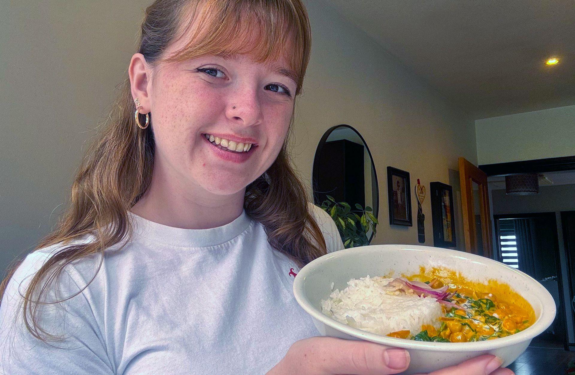 Mia with light brown hair with a fringe wearing a white t shirt is smiling holding a bowl of chickpea curry.
