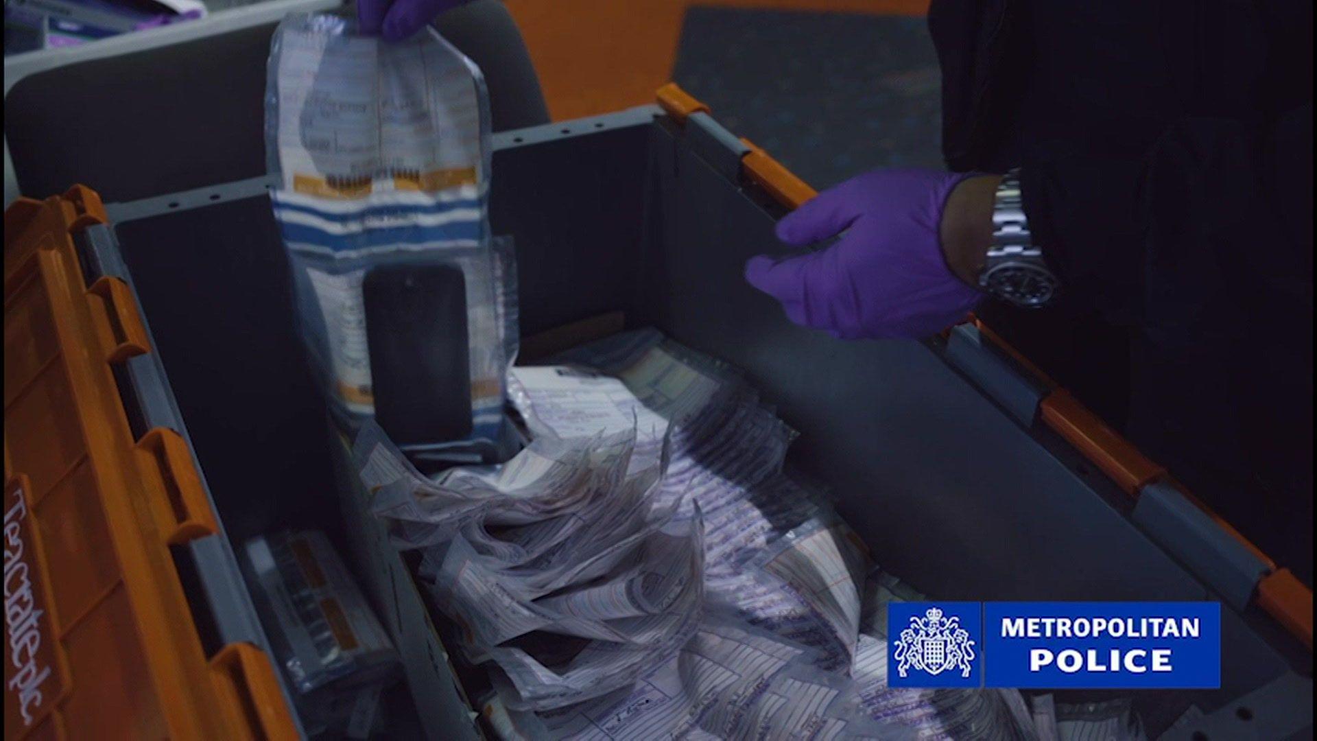 Police footage shows an individual wearing purple latex gloves picking up an evidence bag containing a phone. Beneath it, there appear to be many other evidence bags inside a large grey and orange container.