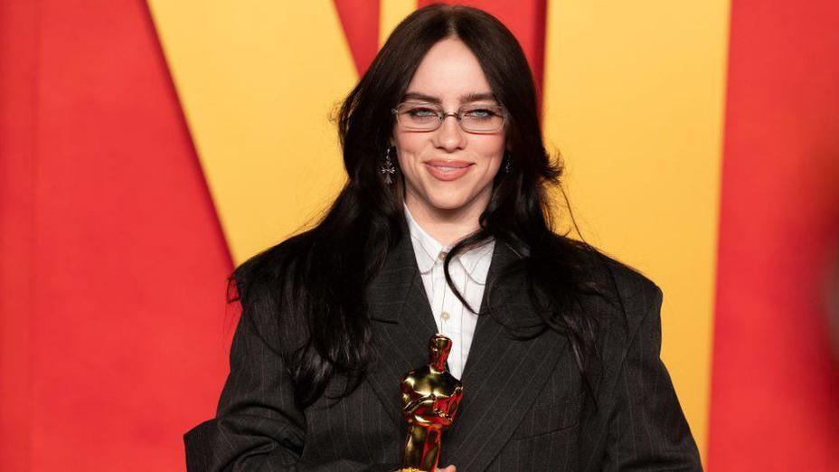 Billie Eilish, wearing a black suit and white shirt, holding her Oscar award at the 2024 Vanity Fair Oscar After Party