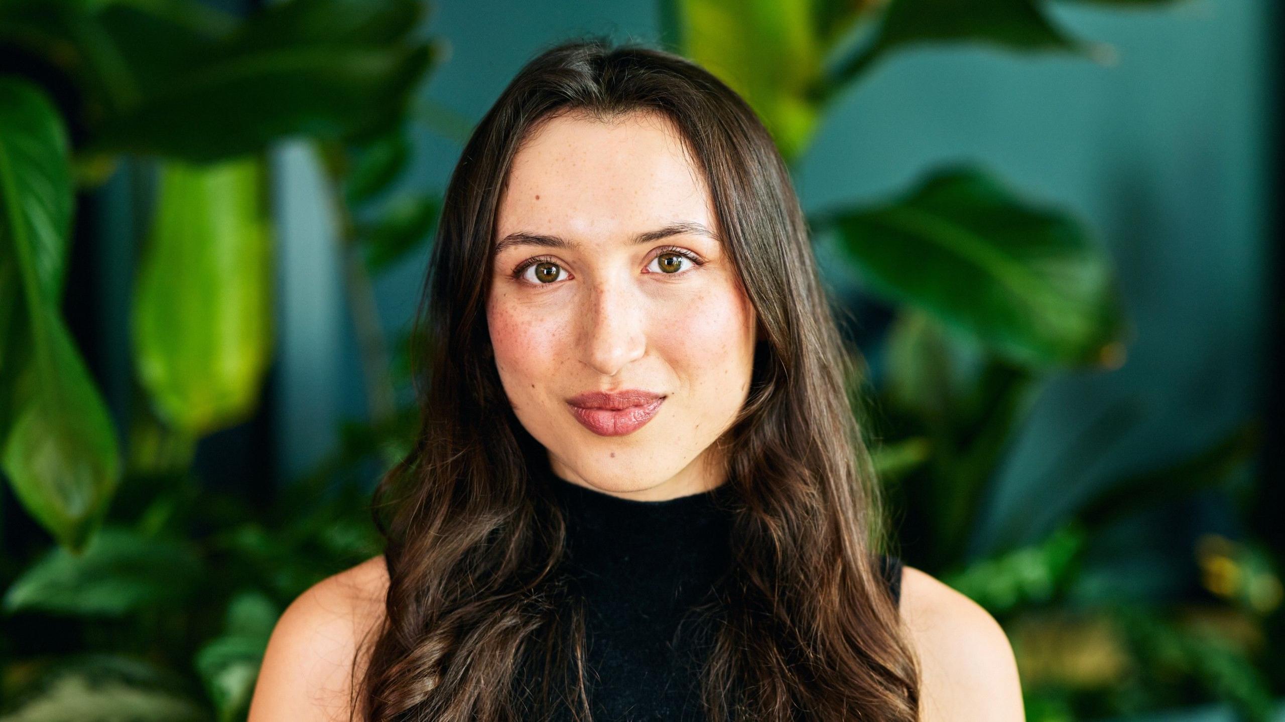 Yumi Liberman smiles at the camera, wearing a black, sleeveless top, and green plants behind her