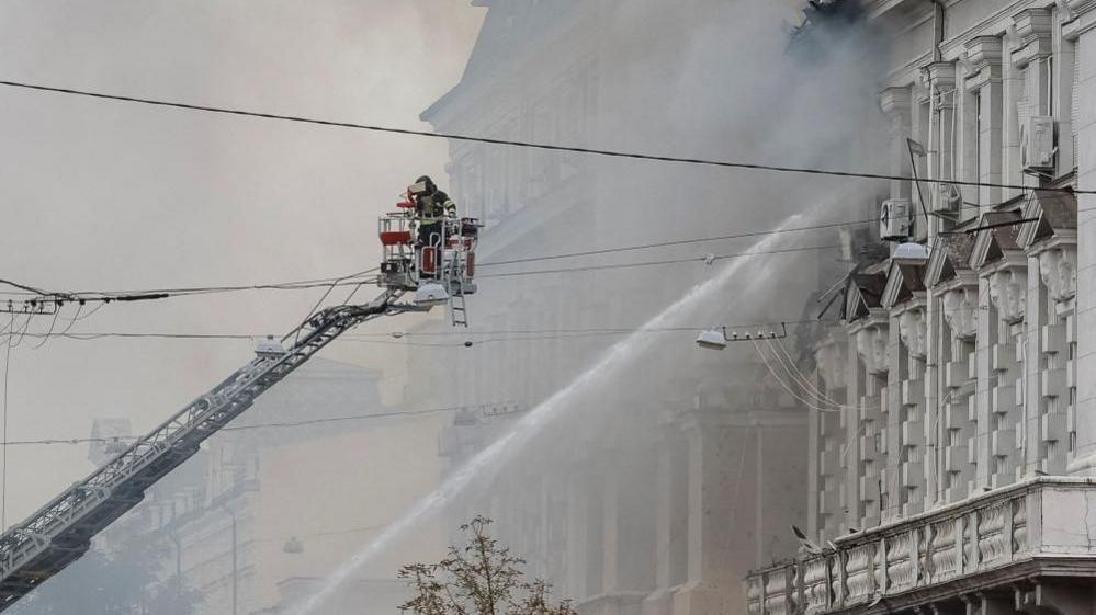 A firefighter on an extended platform helps with hosing down the building