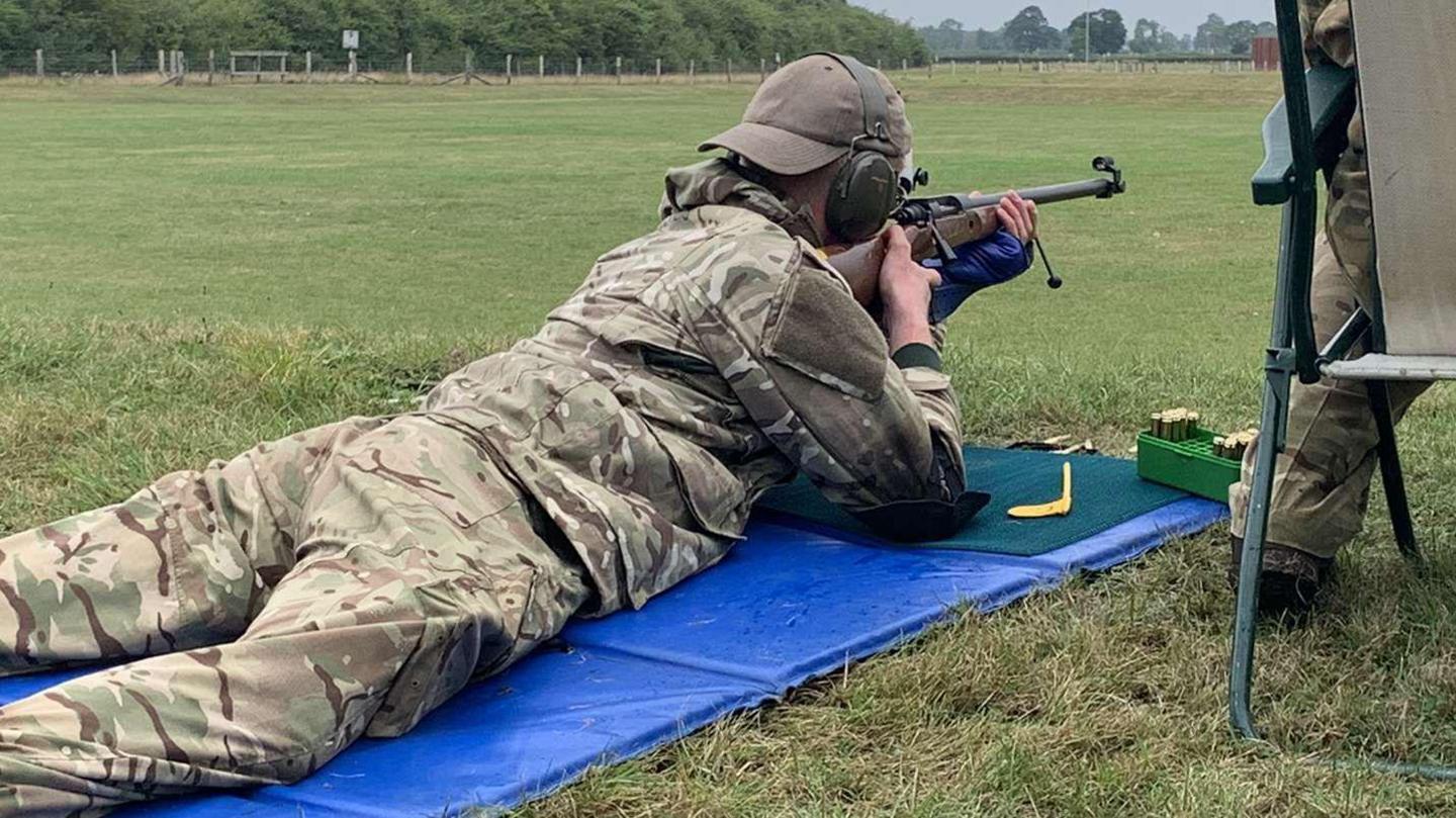 Jackson pictured when he was in the cadets. He is wearing a camouflage-patterned uniform and a black military beret. He is holding a rifle while laying on the floor. He is looking down the scope.
