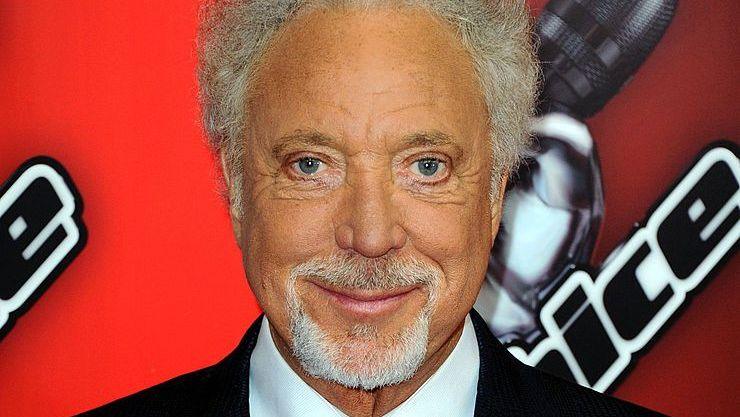 Sir Tom Jones in a blue suit stood in front of a red backdrop with the words The Voice UK on it.