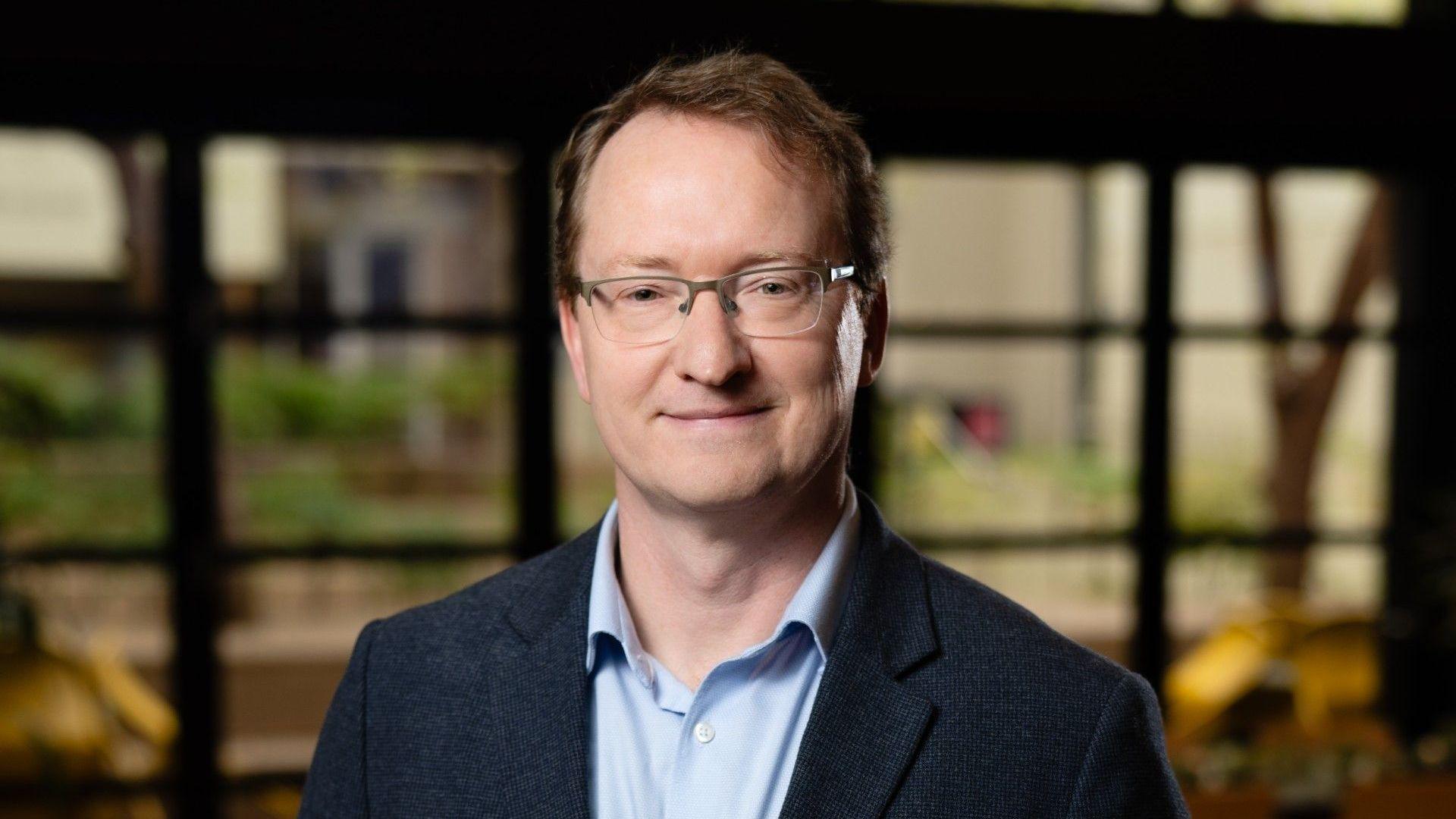 A middle aged man with short brown hair and glasses wearing a blue shirt and jacket smiles at the camera