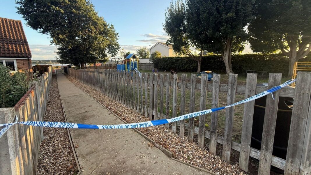 Search continues for boy, 3, missing at playground Blue and white police tape cordoning off a pathway next to a small park, which is lined with trees and a bush.