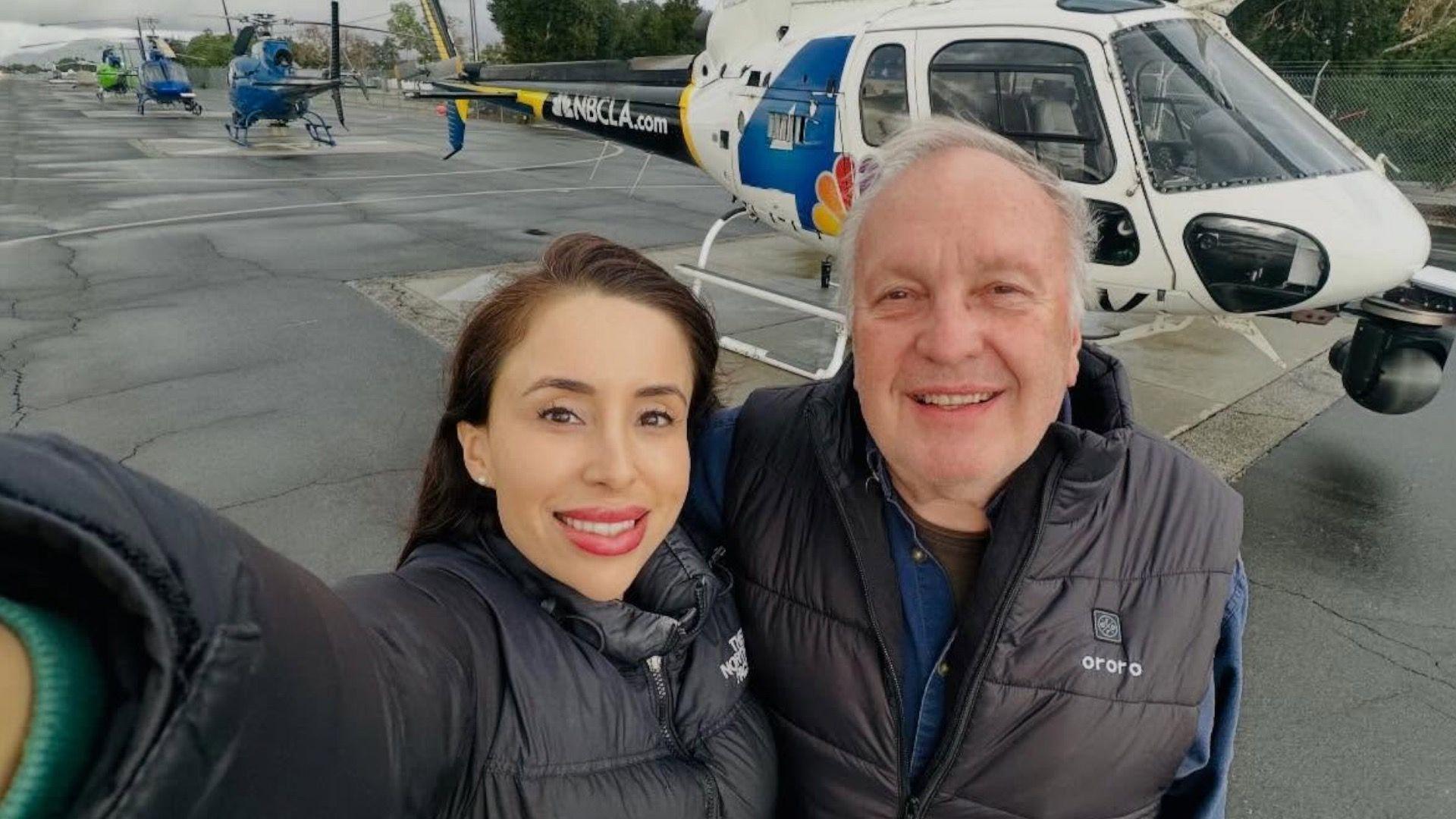 Photojournalist Eliana Moreno and pilot George Marciniw are hown taking a selfie. She is wearing a black puffy jacket and smiling and he is wearing a black puffy vest and smiling. They are standing in front of a helipcopter on the ground