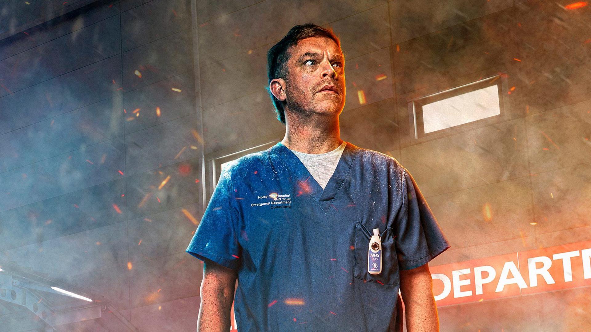 Promotional image of William Beck in character as Dylan Keogh. He is photographed from the waist up and is wearing blue medical scrubs. The set of the emergency department can be seen behind him. His face is covered in soot and embers from a fire can be seen around him. There is smoke surrounding him.