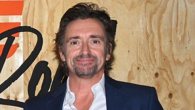 Richard Hammond is photographed from the shoulders up, standing in front of a promotional sign for the TV series 'The Grand Tour'. His face is facing slightly to the side and he is smiling at the camera. He is wearing a black suit jacket and a white shirt, with the top button undone.