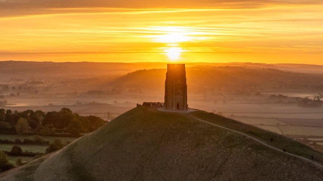The sun rises in a yellow sky over the top of Glastonbury Tor, which is a monument on the top of a hill. People stand at the base of the Tor watching.