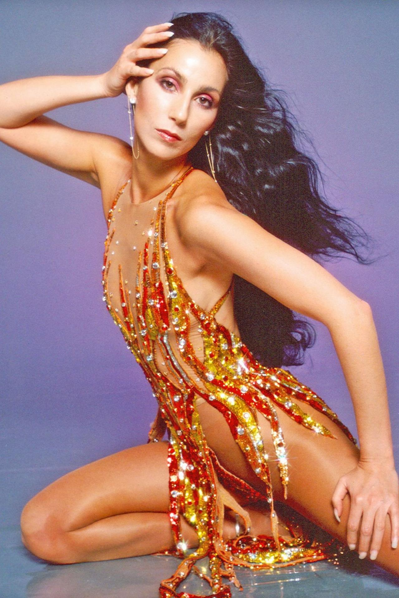 Studio shot of Cher posing in a bright orange and red bejewelled gown, which looks like flames.