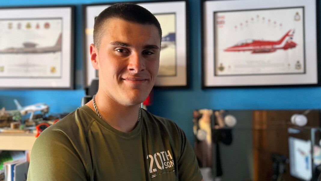 Ben Smith looking into the camera and smiling. He is a young man with dark hair which is shaven. He is wearing a khaki T-shirt with a white slogan and a necklace. Smith is pictured in his bedroom and the walls are covered in framed photos of military aircraft. The walls are painted blue.