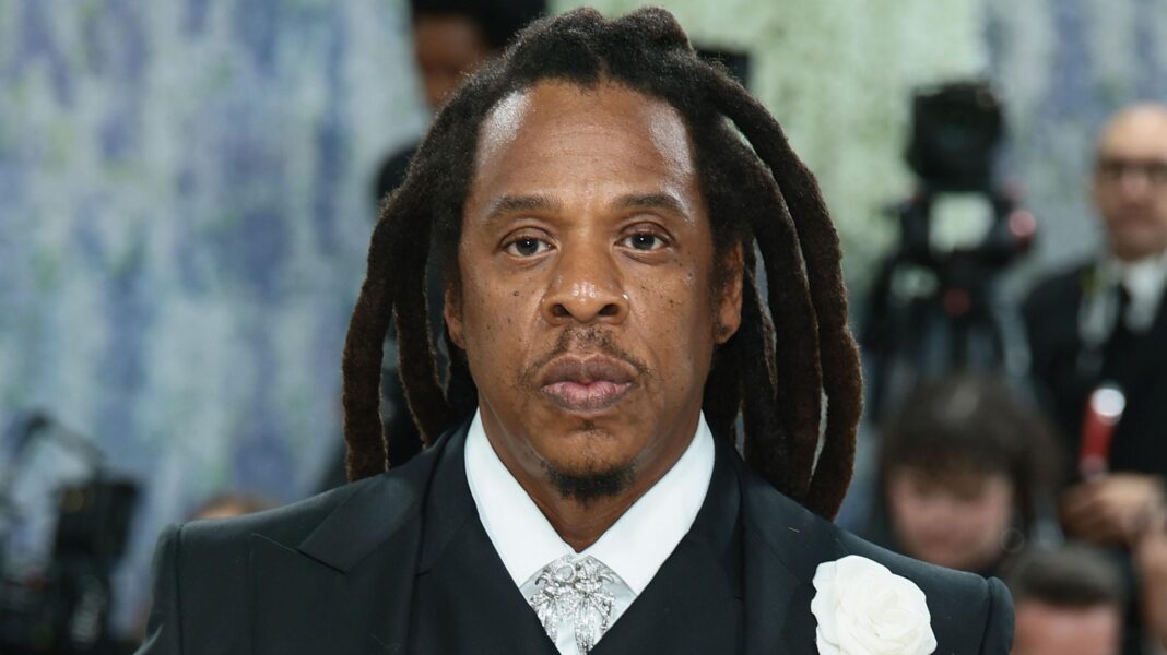 Jay-Z wearing a black suit, white shirt, grey patterned tie and white rose