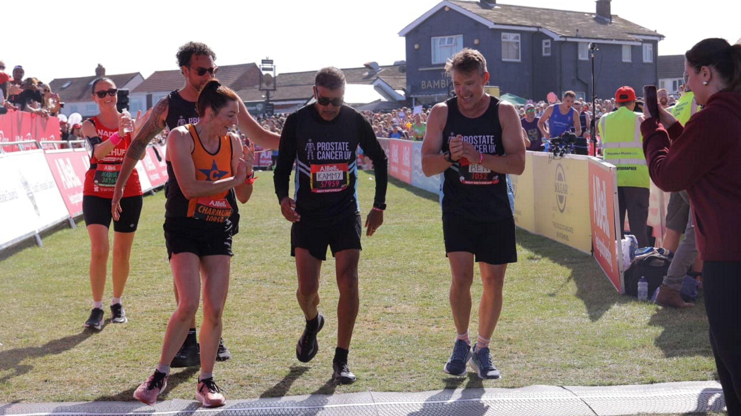 Five runners crossing the finishing line. To the left, and slightly behind, is a woman holding a water bottle, and appearing to be clapping her hands. A woman in front, wearing an orange vest, is also applauding. Behind her is a man who appears to be patting the centre runner, who is football personality Chris Kamara, on the back. He is wearing a black t-shirt with Prostate Cancer UK and dark glasses. Next to him another man, also wearing a black Prostate Cancer t-shirt, is clapping. Crowds of spectators line the hoardings behind the finish line.