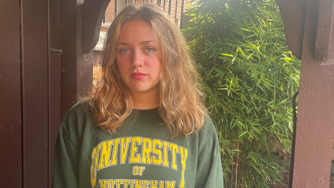 Woman with blonde hair and blue eyes stood in front of bush wearing a University of Nottingham jumper