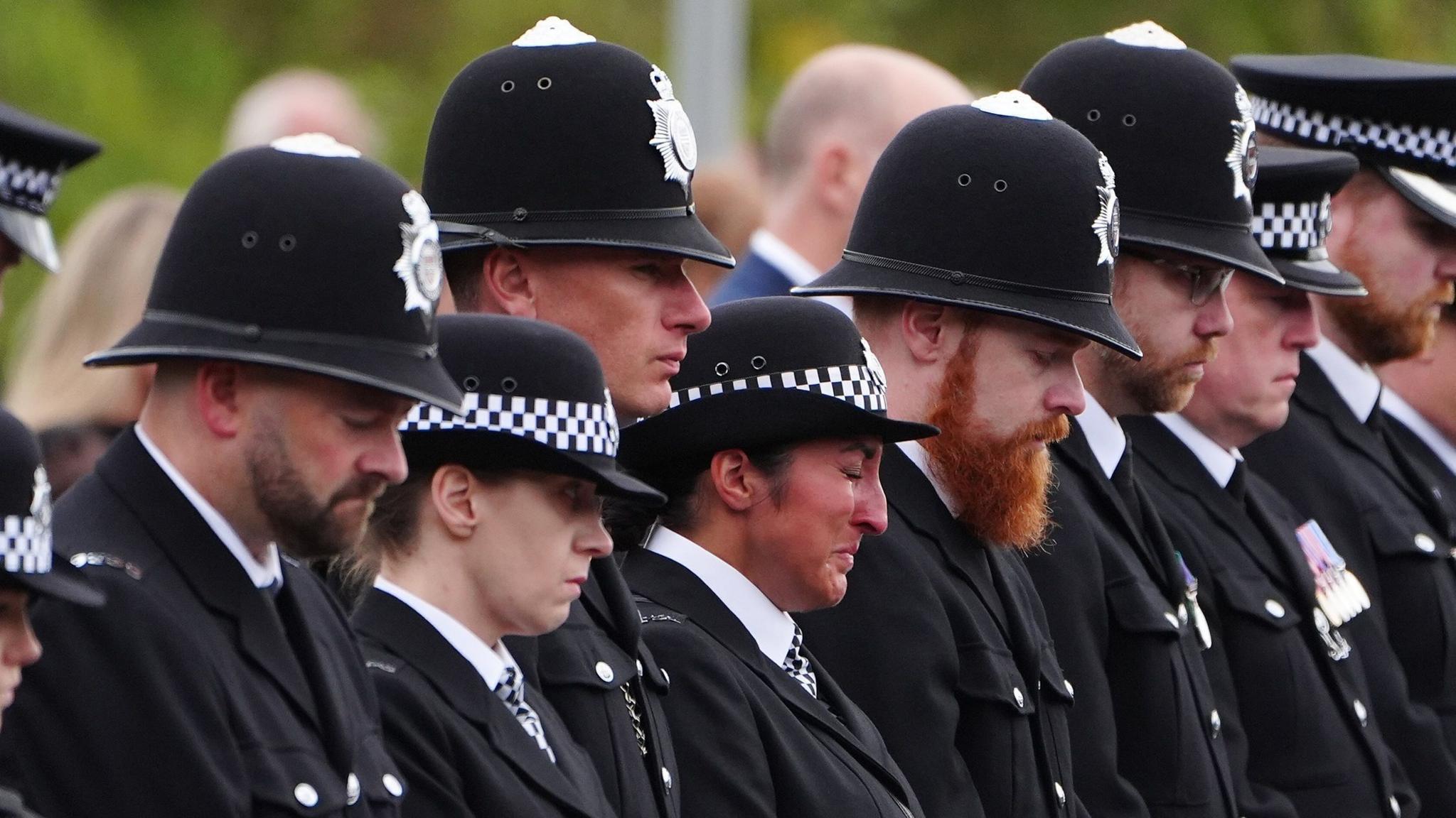 A line of police officers in all black uniform looking emotional with their heads bowed. There are six male officers and two female, some wearing peaked caps and others custodian helmets. There is a woman in the centre of the line who looks more emotional than the others and has her eyes closed while crying.
