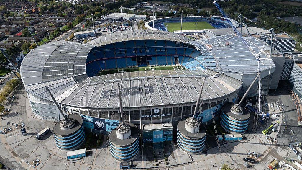 An aerial view shows The Etihad Stadium, the home ground of Manchester City football club, with the Manchester skyline in the background.