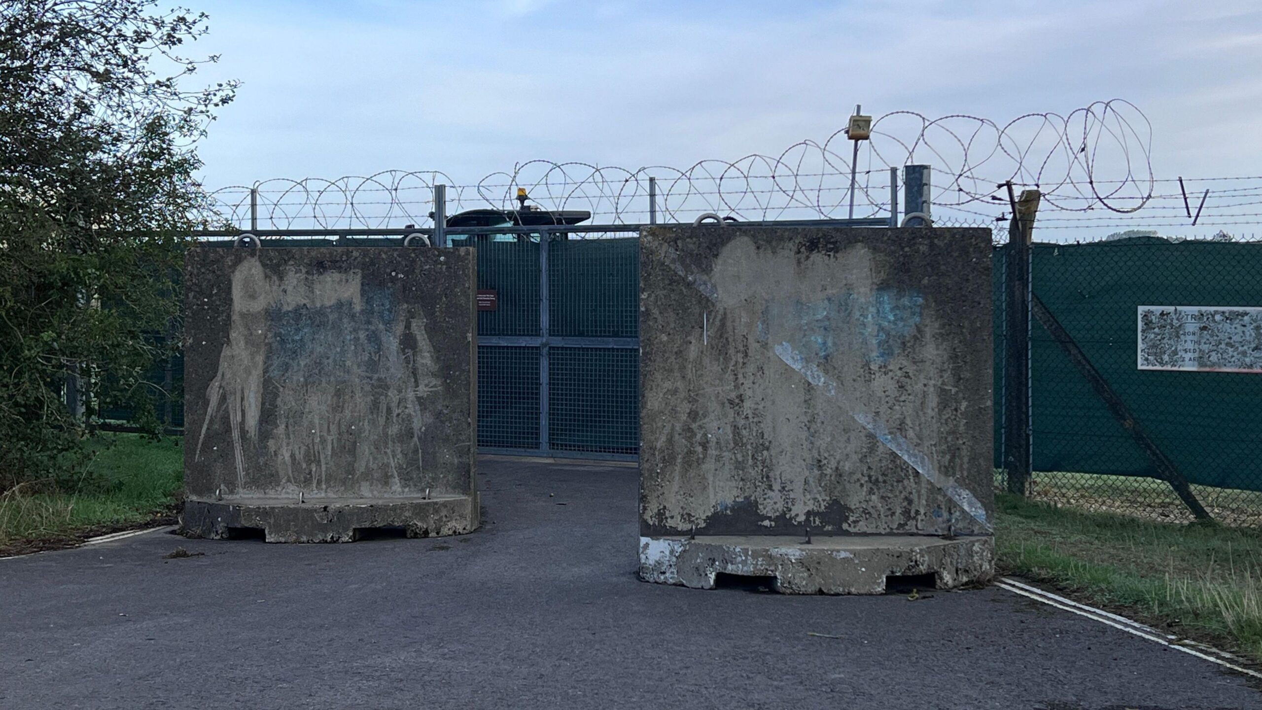 Large concrete barriers positioned in front of a metal gate at RAF Fairford. The fence is covered with mesh to prevent onlookers, and is topped with coiled barbed wire. Behind the fence is a large truck parked directly in front of the entrance.