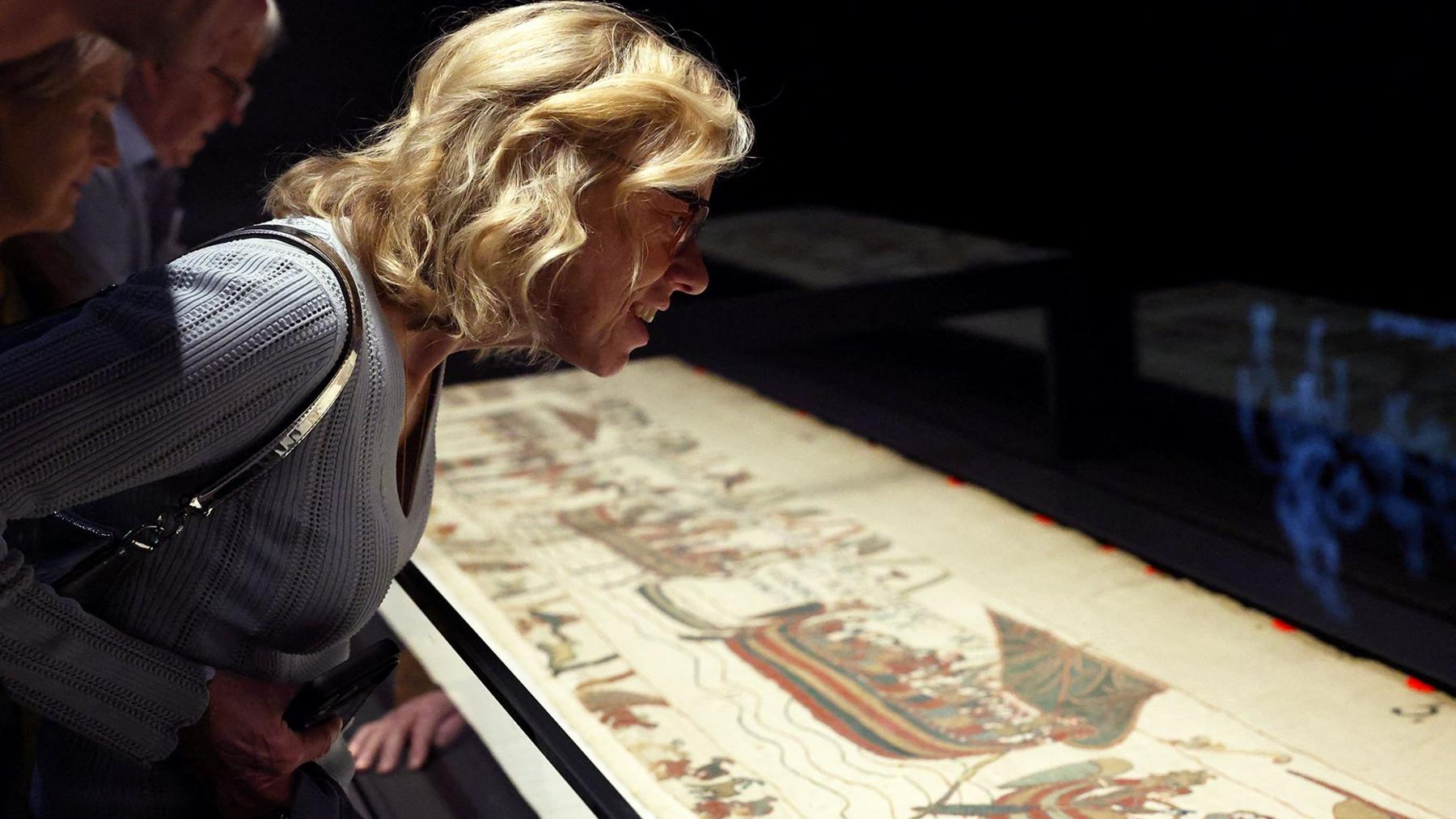A woman with blonde hair and glasses looking closely at the tapestry