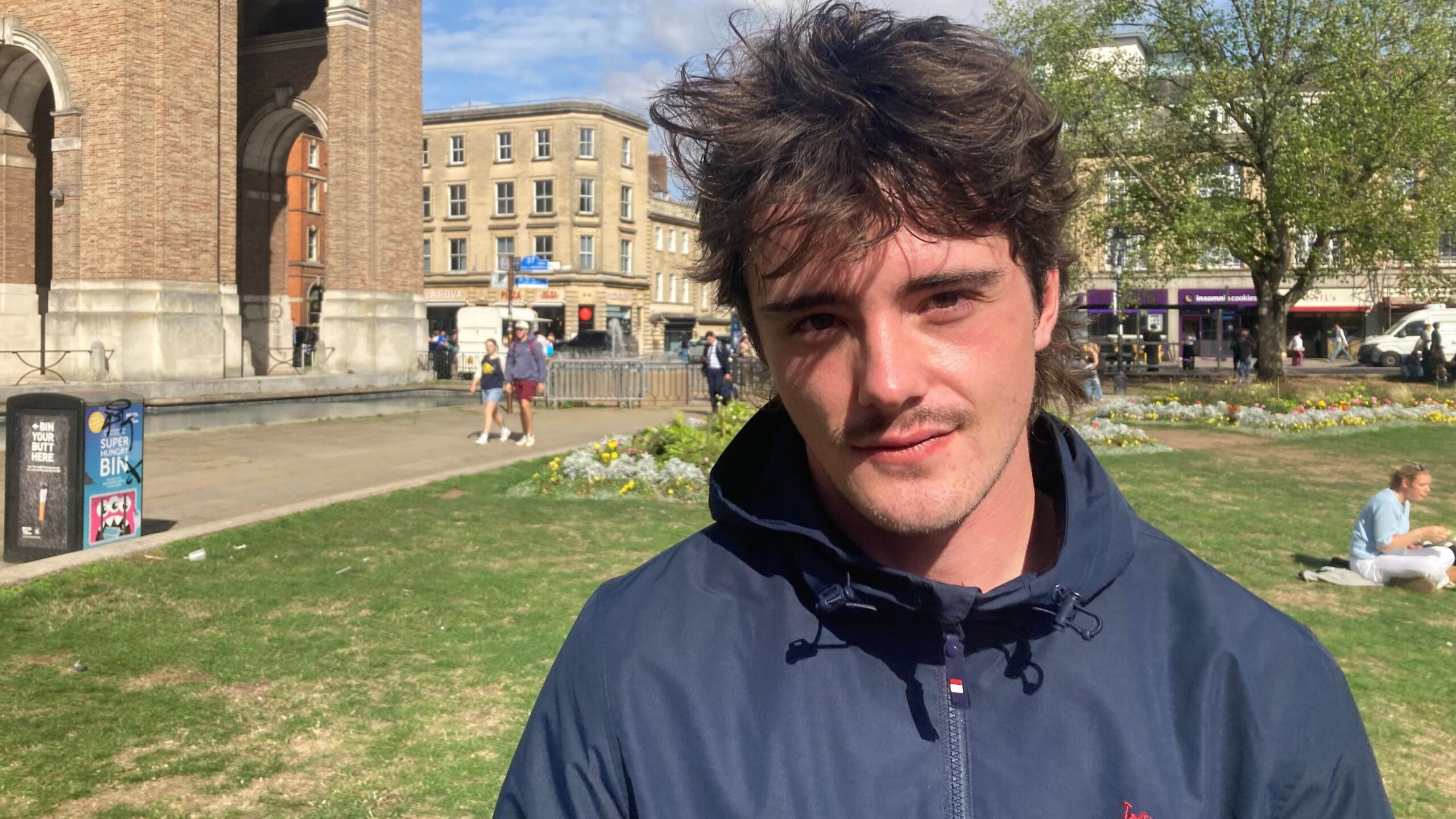 Jamie Crook wearing a blue zip-up windbreaker jacket and looking at the camera. He has brown eyes and dark brown hair styled into a mullet. It is a bright sunny day, and in the background there are people sitting on the grass at College Green.