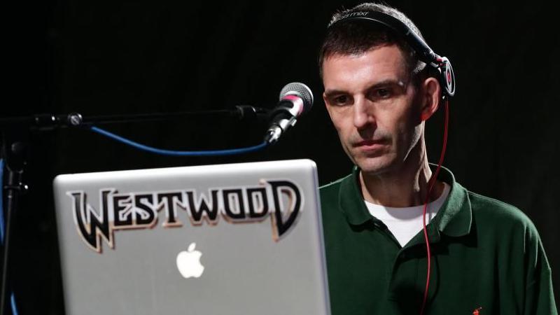 Tim Westwood performing on stage. He is wearing headphones and has a microphone and a computer screen in front of him. The background is black.