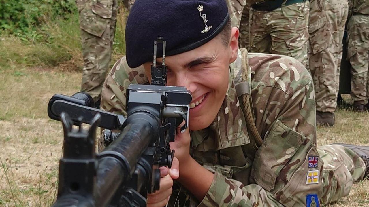 Smith pictured when he was in the cadets. He is wearing a camouflage-patterned uniform and a black military beret. He is holding a rifle while laying on his front on the floor. He is smiling as he looks down the scope.