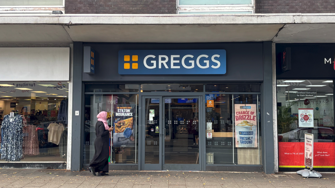 Image shows Greggs - a blue and yellow sign above a shopfront with glass doors. A woman with a pink headscarf is walking past.