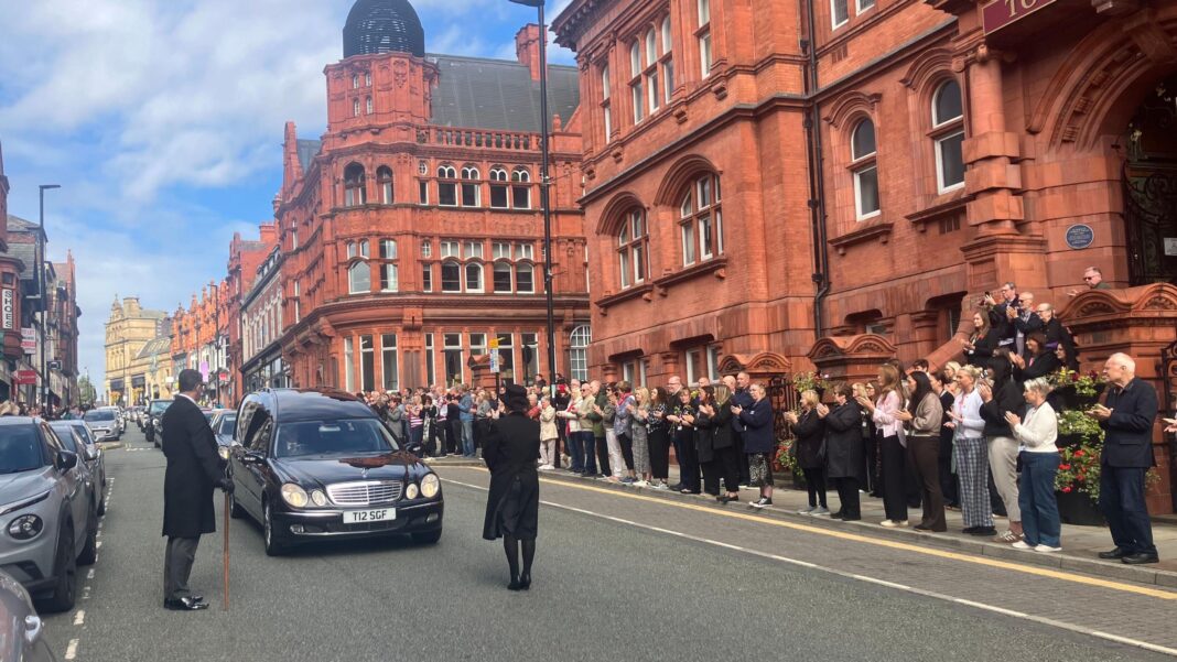 Crowds of people watch the funeral procession drive though a town centre. A black Mercades leads the procession, with two men in black suits leading the convoy.